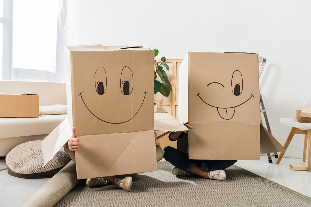 Two people sitting on the floor with cardboard boxes on their heads, looking excited.